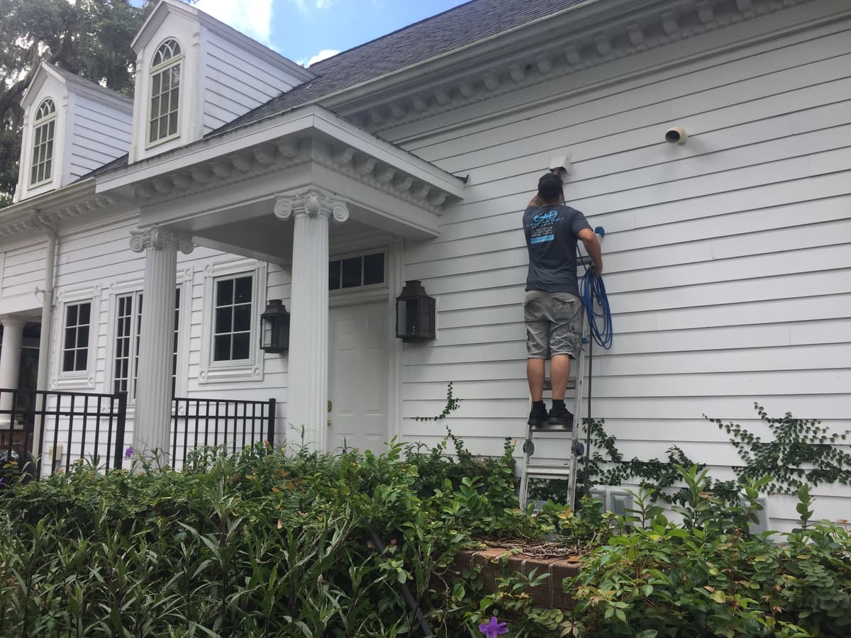 A technician on a ladder cleaning an exterior dryer vent on a two-story home