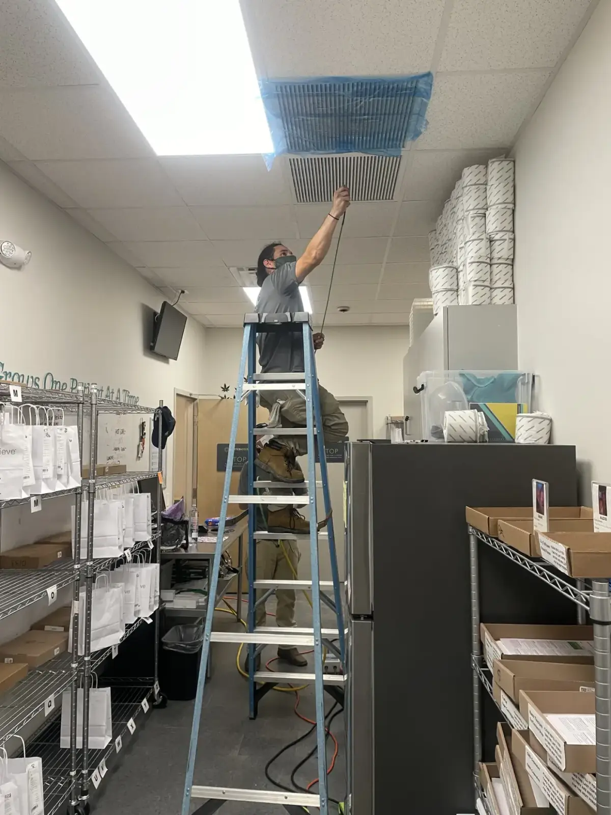 A technician on a ladder cleaning a vent in a commercial storage room