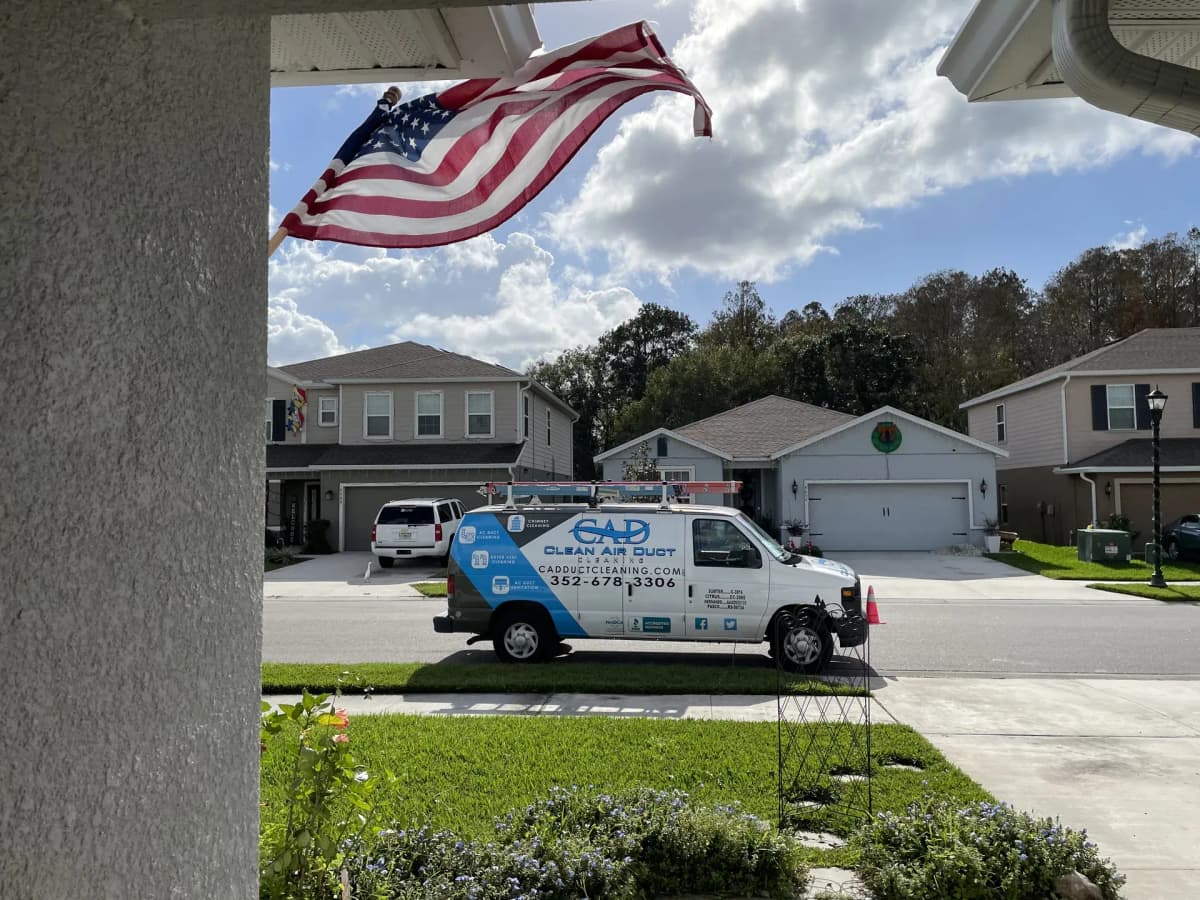 A Clean Air Duct Cleaning van parked at the kerb outside a single-storey Florida home, seen from the front porch with a US flag overhead