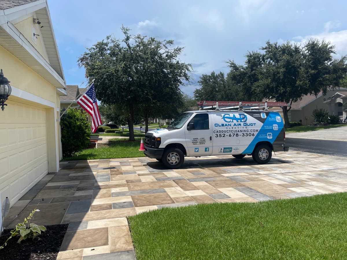 A Clean Air Duct Cleaning van in the driveway of a single-story home with an American flag by the door