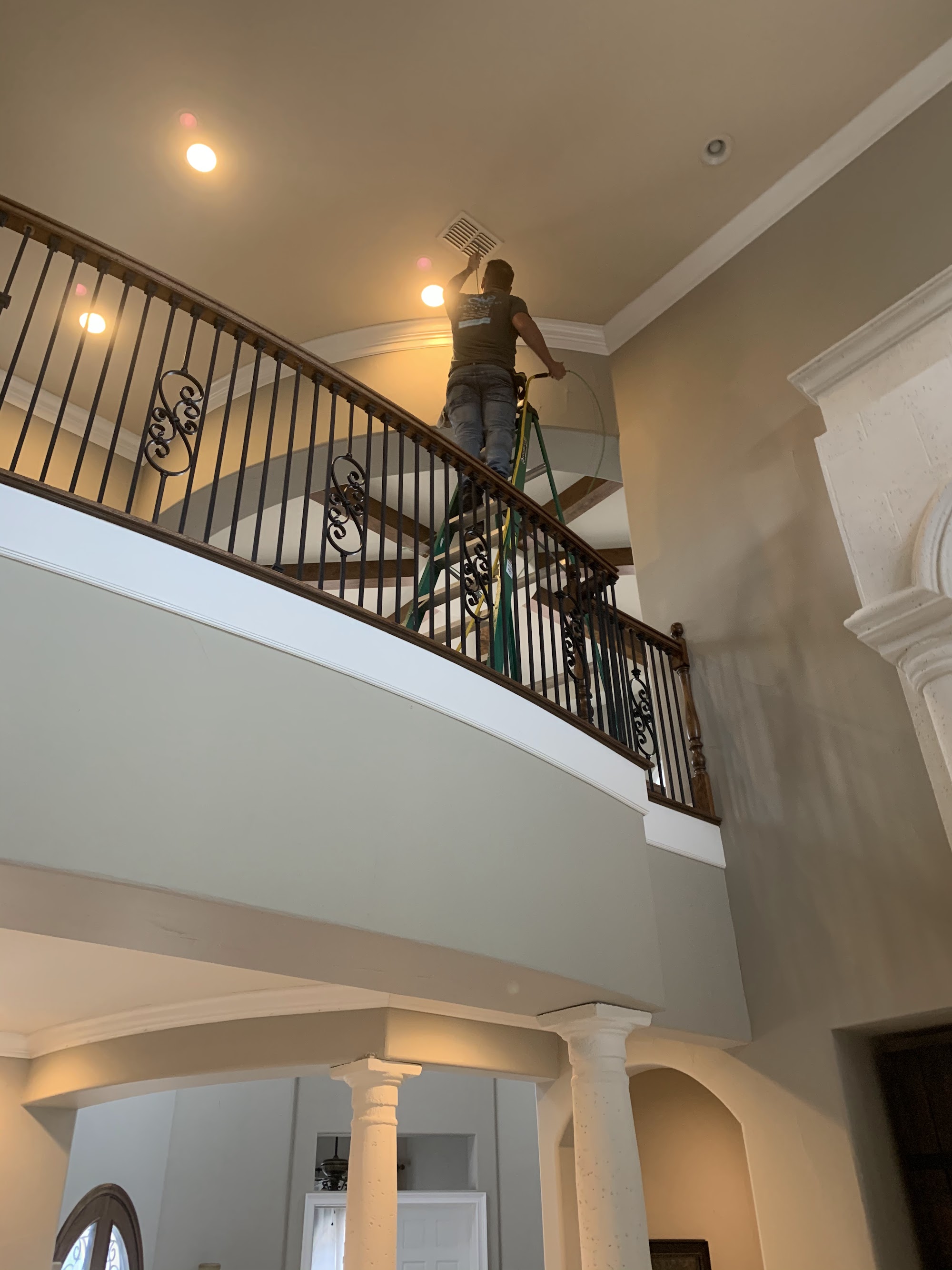 A Clean Air Duct Cleaning technician on a tall ladder cleaning a return vent in a two-storey Florida foyer